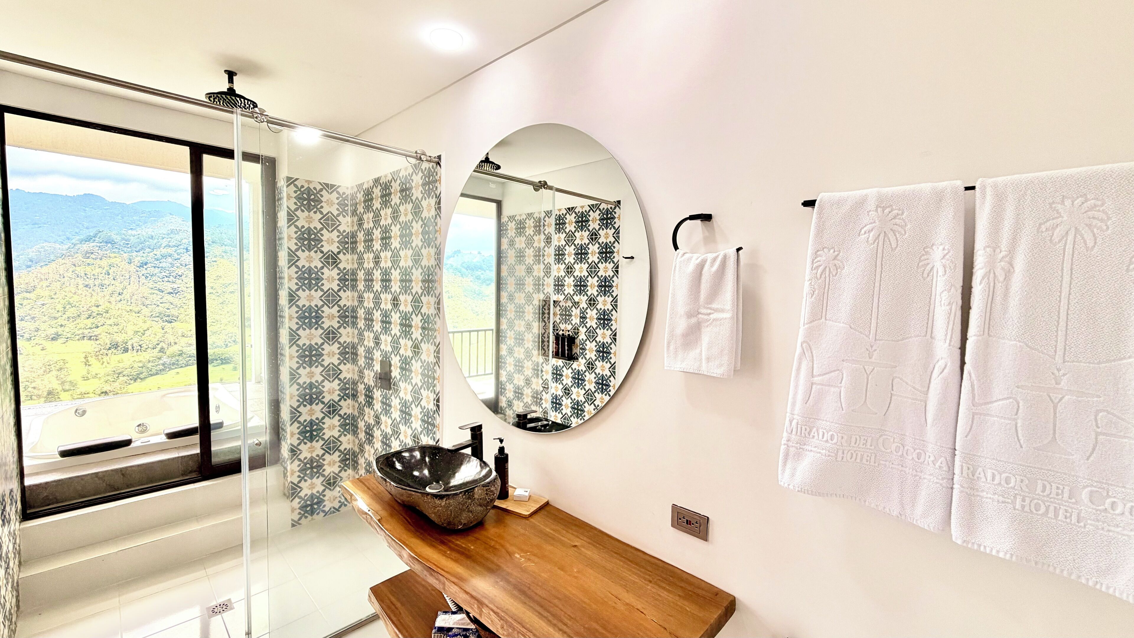 A bathroom with a large mirror, a wooden vanity, a sink with a large bowl, patterned shower curtains, and two white towels on a rack.