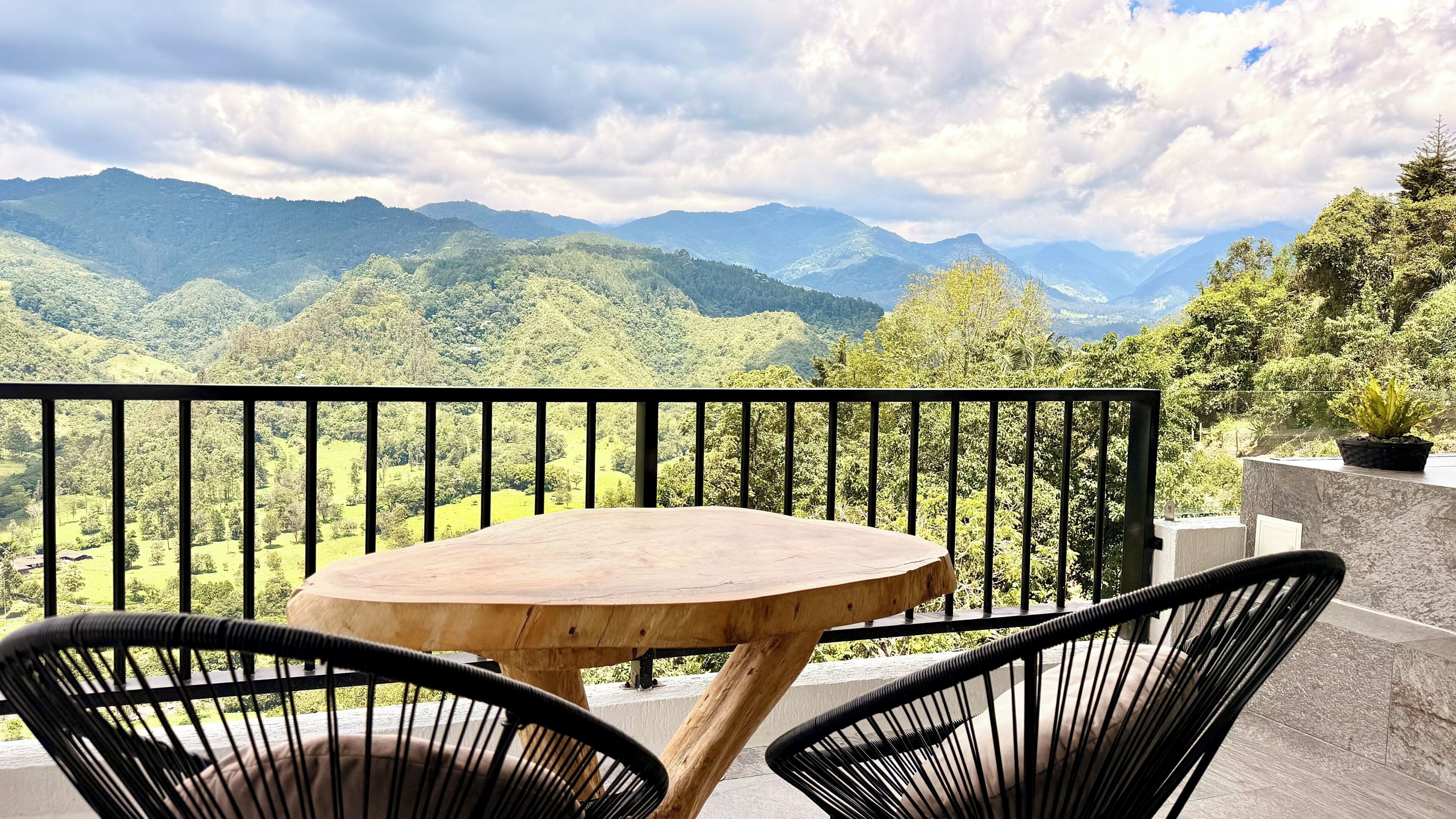Une table ronde en bois, accompagnée de deux chaises noires, est placée sur un balcon doté d’une balustrade noire, d’où l’on peut admirer un paysage montagneux.