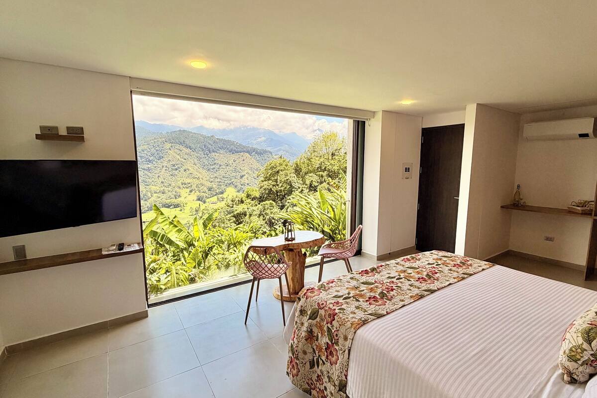A bedroom with a bed, a TV, a table with chairs, and a view of a mountainous landscape.