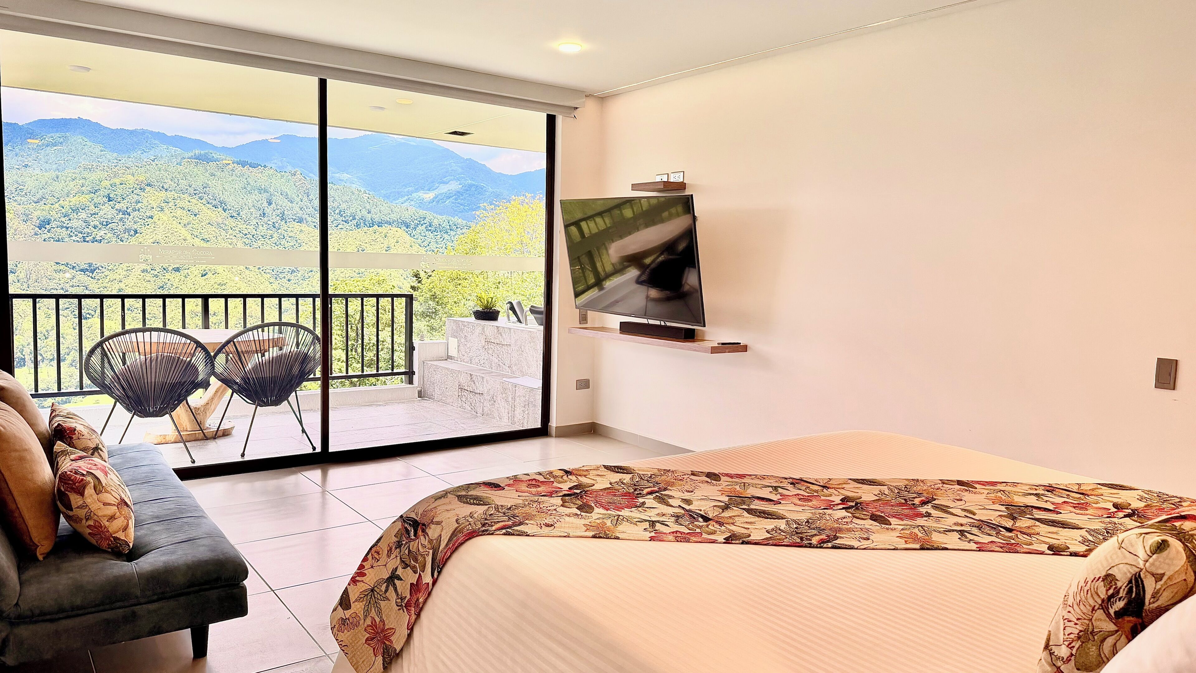 A hotel room with a balcony, a bed, a sofa, and a TV.