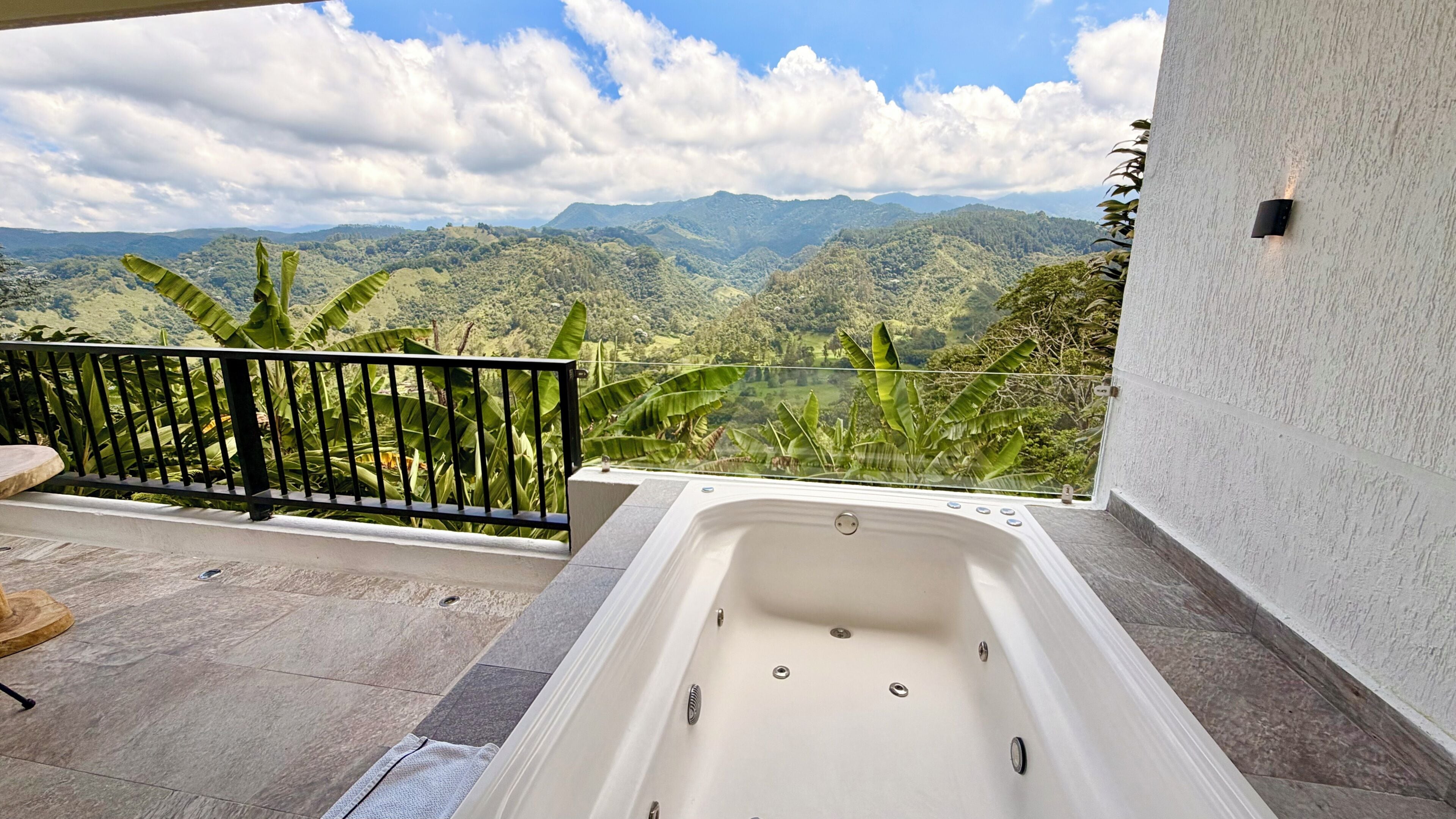 A balcony with a view of mountains and greenery, a bathtub, and a small table.