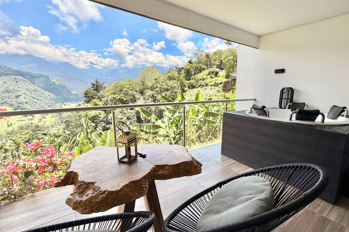 A balcony with a view of mountains and greenery, a wooden table, two wicker chairs, and a modern desk with a chair.
