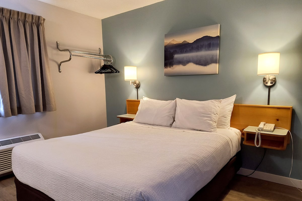 A hotel room with a large bed, a bedside table with a lamp, a painting on the wall, and a window with curtains.
