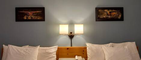 A hotel room with two beds, a wooden headboard, a bedside table with a lamp, and two framed pictures on the wall.