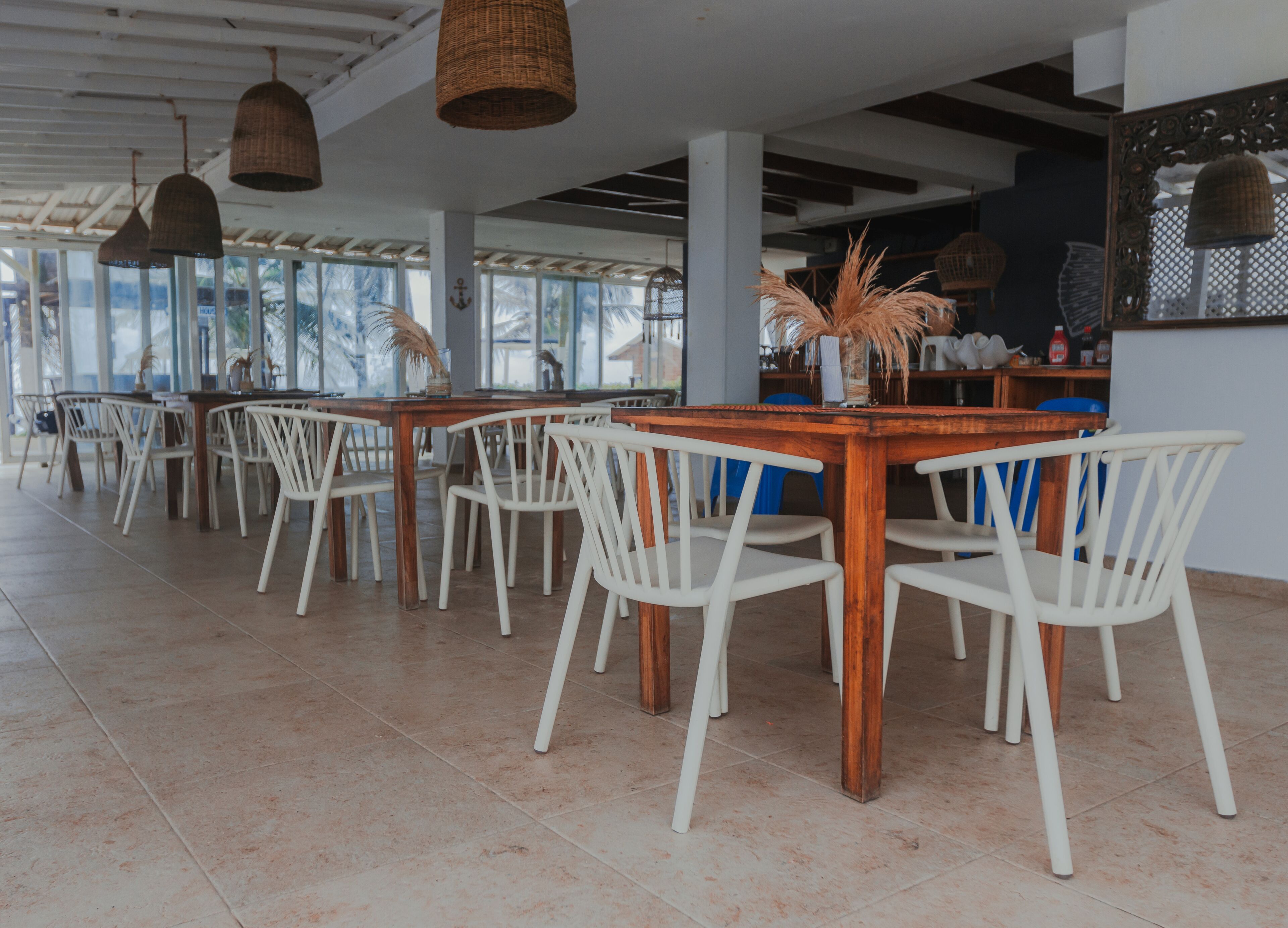 Uma ampla sala de jantar com cadeiras brancas e mesas de madeira, janelas grandes e luminárias de vime suspensas.