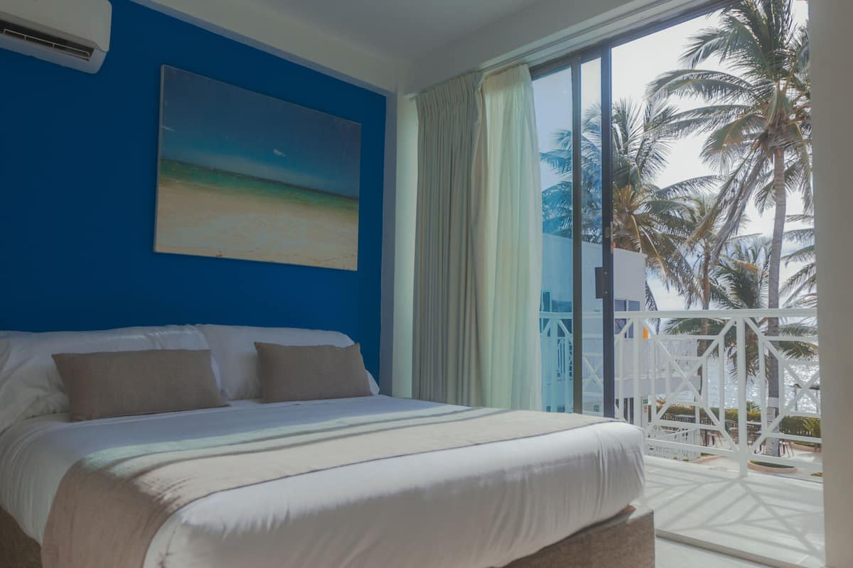 A hotel room with a large bed, a painting of a beach, and a view of palm trees through the window.