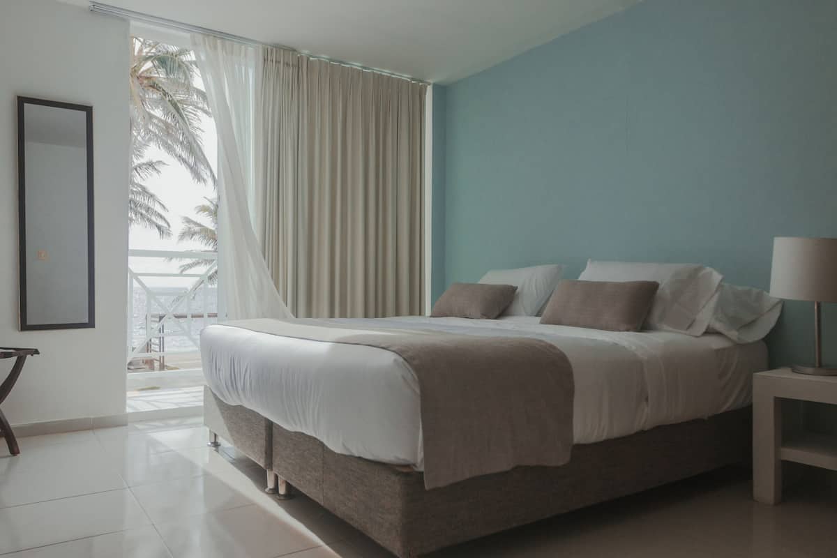 A bedroom with a bed, a nightstand, a lamp, and a view of palm trees through a window.