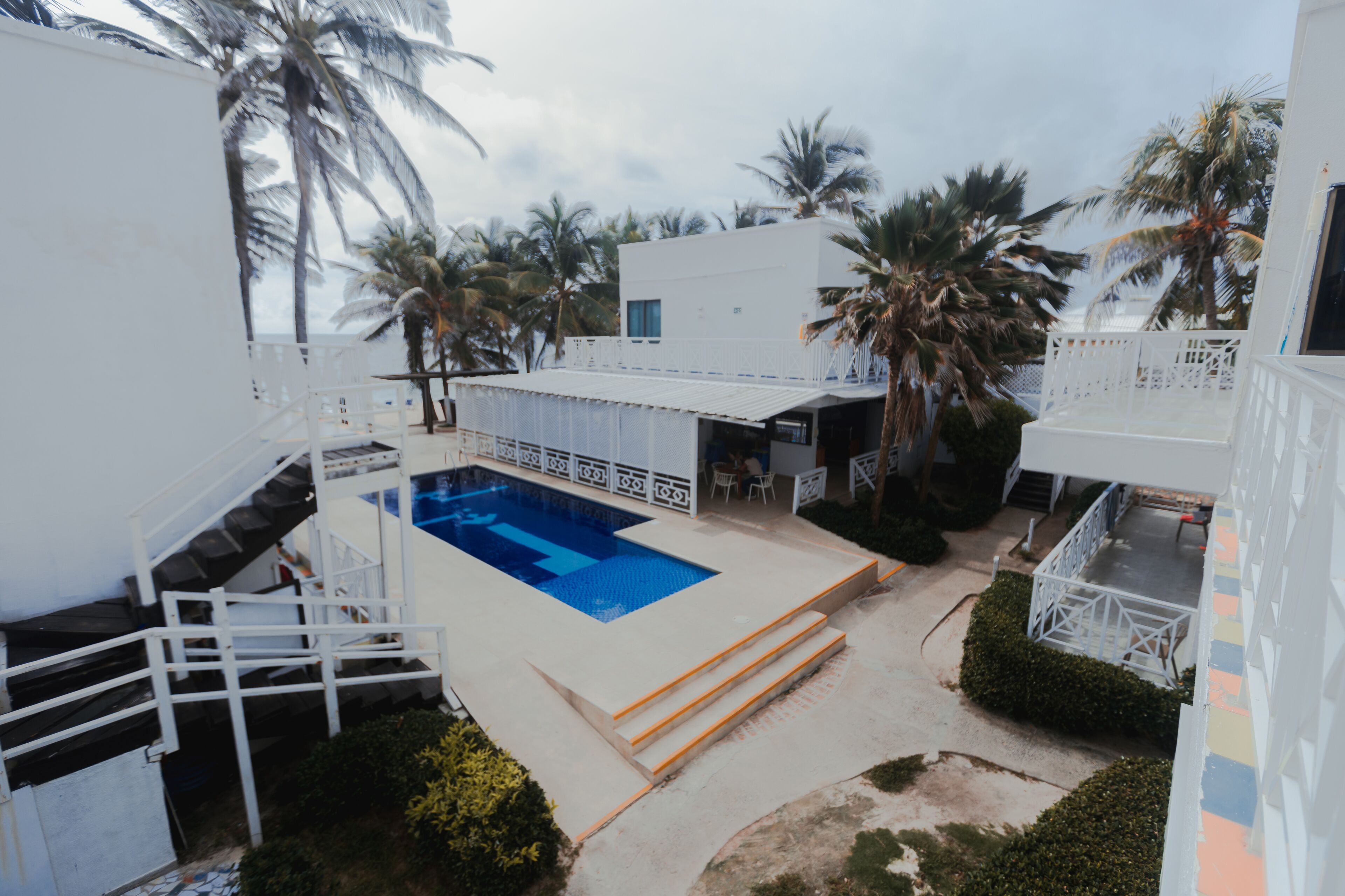 Uma área de piscina com escadas dando acesso à piscina, cercada por palmeiras e edifícios brancos.