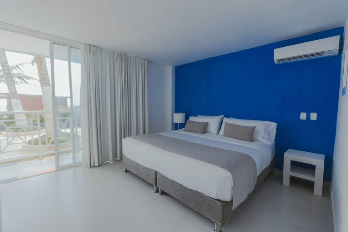 A modern hotel room with a large bed, a small bedside table, a ceiling air conditioner, and a view of palm trees through the large sliding glass door.