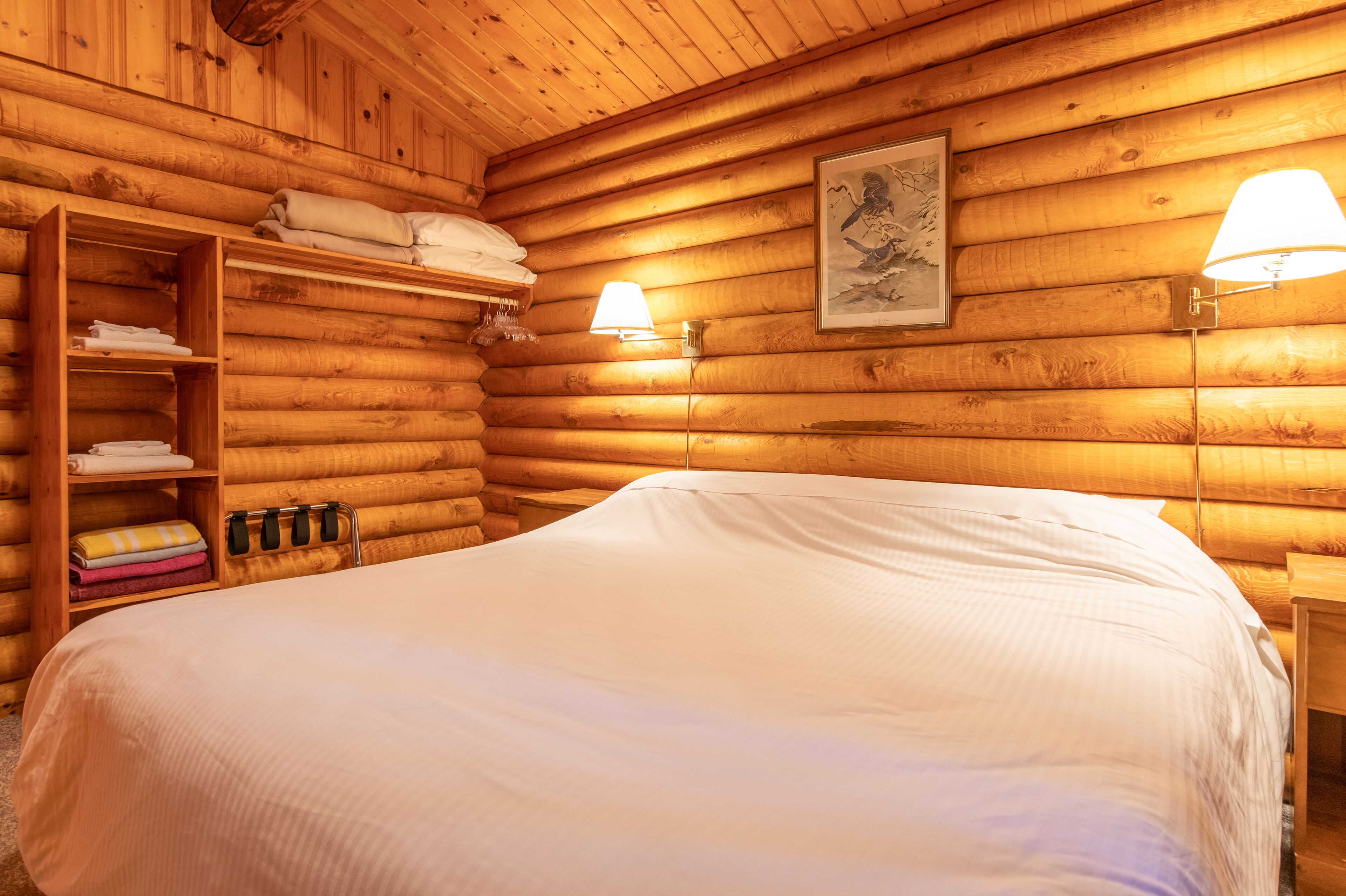 A log cabin-style room with a large bed, wooden shelves with towels, a wall-mounted lamp, and a framed picture.