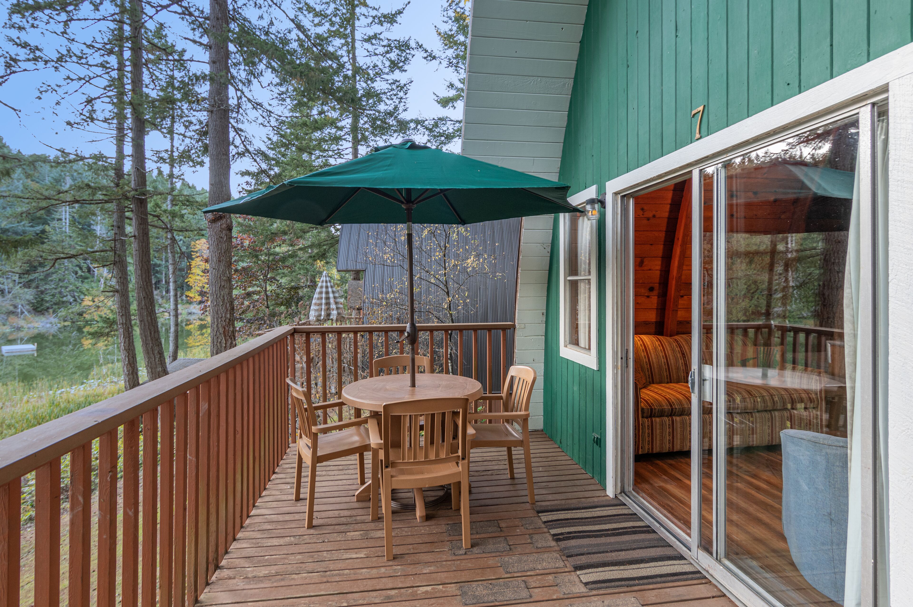 A wooden deck with a table and chairs, a green umbrella, and a sliding glass door leading to a covered seating area.