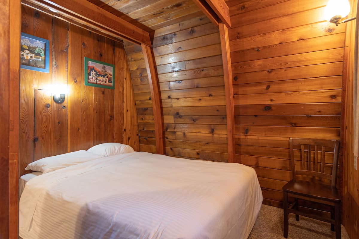 Quarto com painéis de madeira, uma cama, uma cadeira e duas luzes fixadas na parede.
