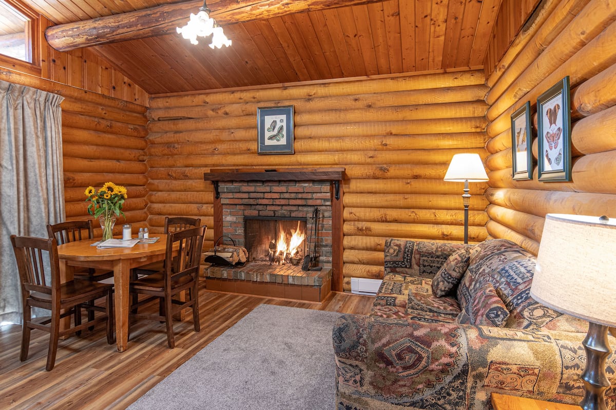 Interior de uma cabana de toras com lareira, móveis de madeira e quadros nas paredes.