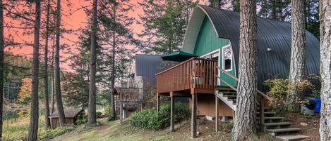 Une cabane à l’extérieur vert et dotée d’une terrasse en bois, entourée de grands arbres et d’un petit étang.