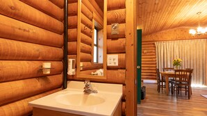A wooden cabin interior with a bathroom featuring a sink, mirror, and towel rack. A dining area with a table and chairs is visible in the background. - Cusheon Lake Resort (Salt Spring Island)