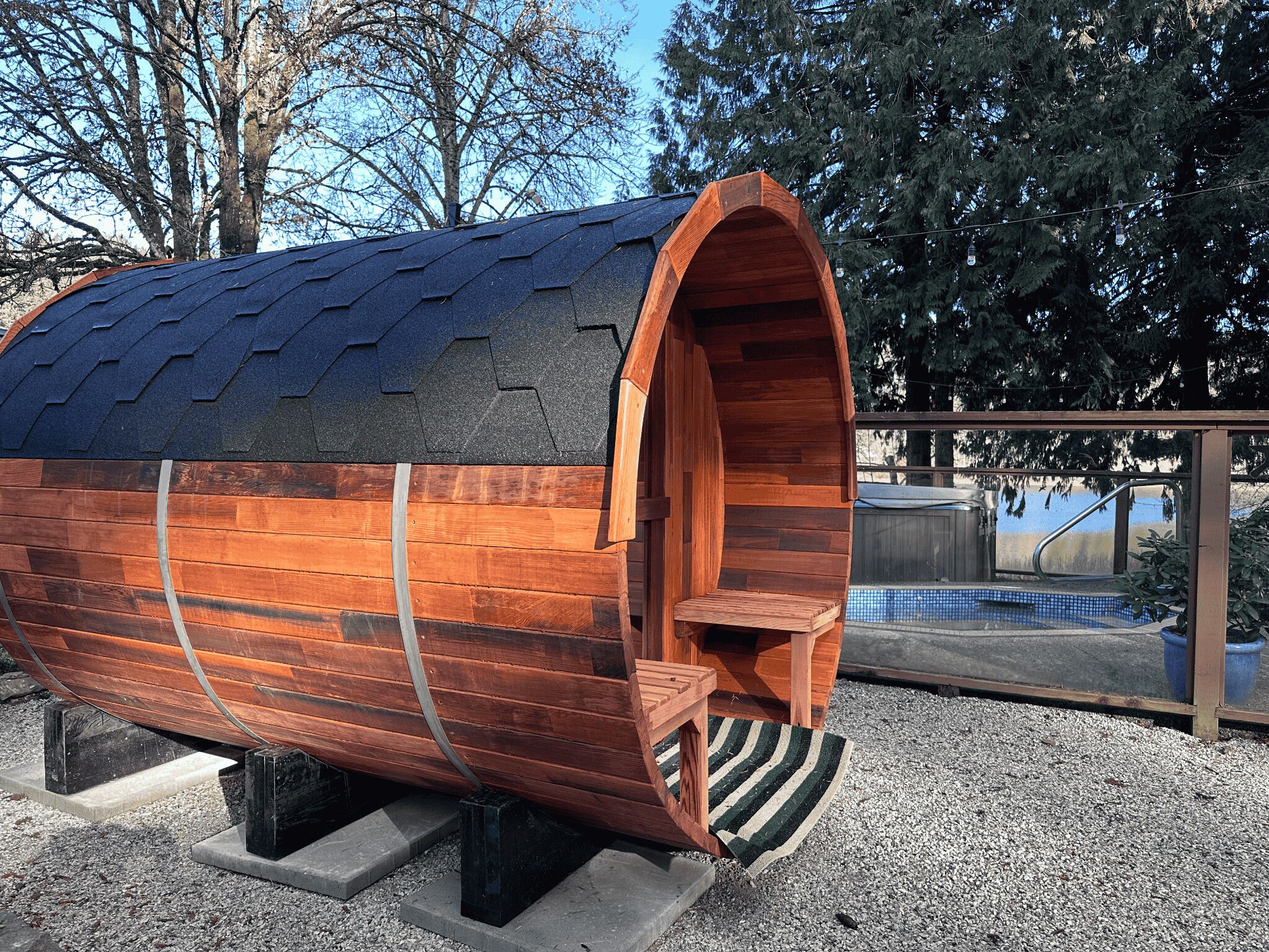 Una estructura con forma de barril de madera, techo metálico, un pequeño banco de madera y barandilla metálica.