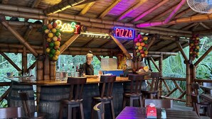 Una zona de bar rústica de madera con un letrero de neón que dice 'BAR' y 'PIZZA', rodeada de sillas y mesas de madera.