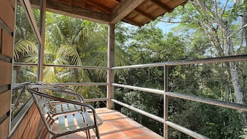 Un espacio exterior cubierto con una silla de metal, baldosas de terracota y una barandilla metálica con vistas a un paisaje verde exuberante.