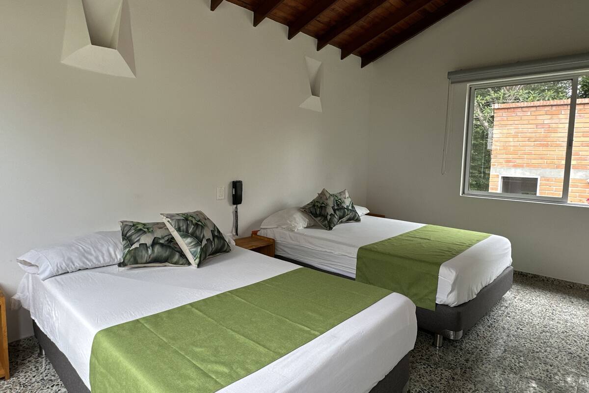 Een kamer met twee bedden, elk met een groene spreidek en witte lakens. Er is een houten hoofdeinde, een nachtkastje met een telefoon, en een raam met uitzicht op een bakstenen gebouw.