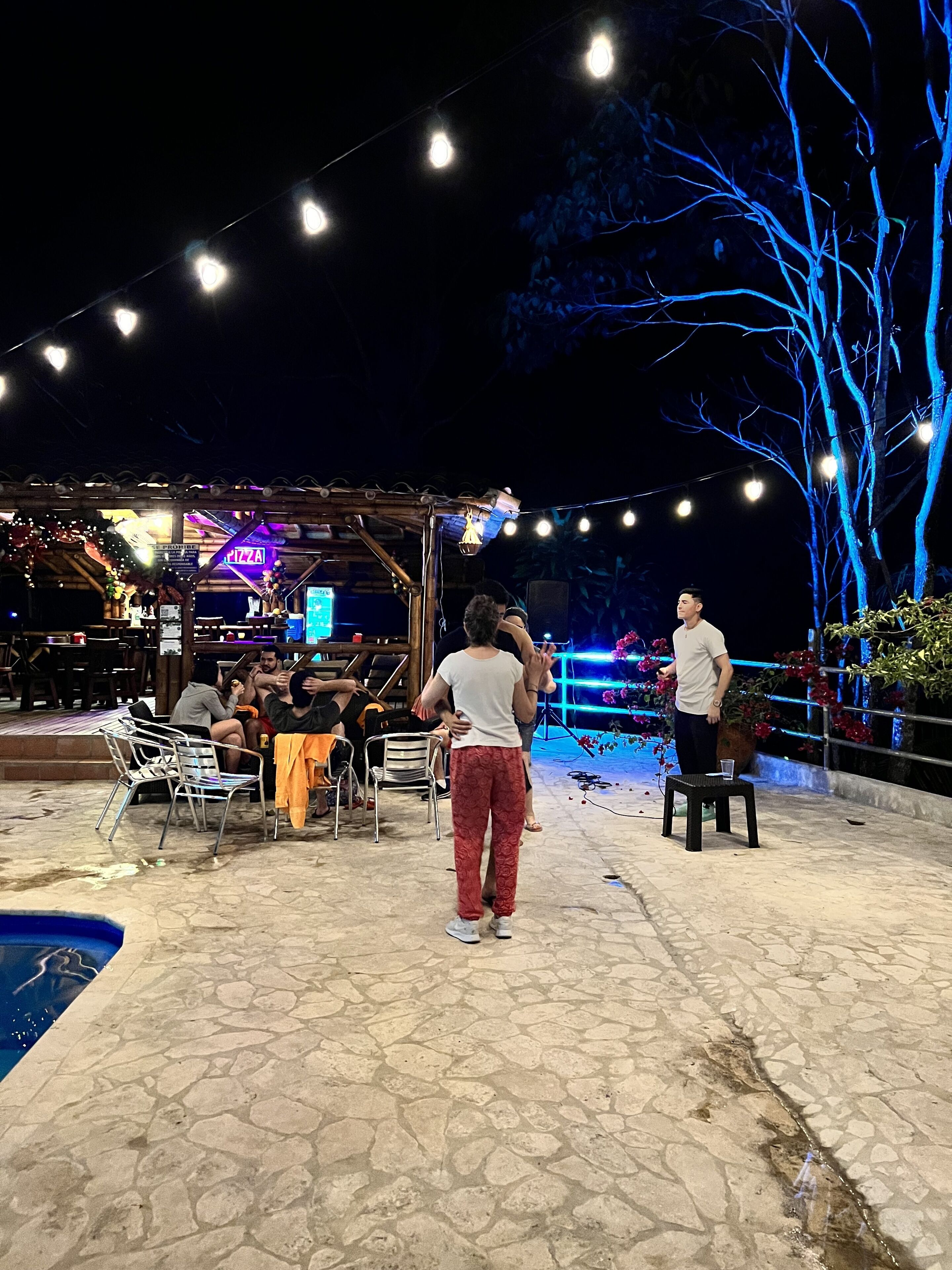 Un patio al aire libre con luces colgantes, una piscina y una zona de estar cubierta.