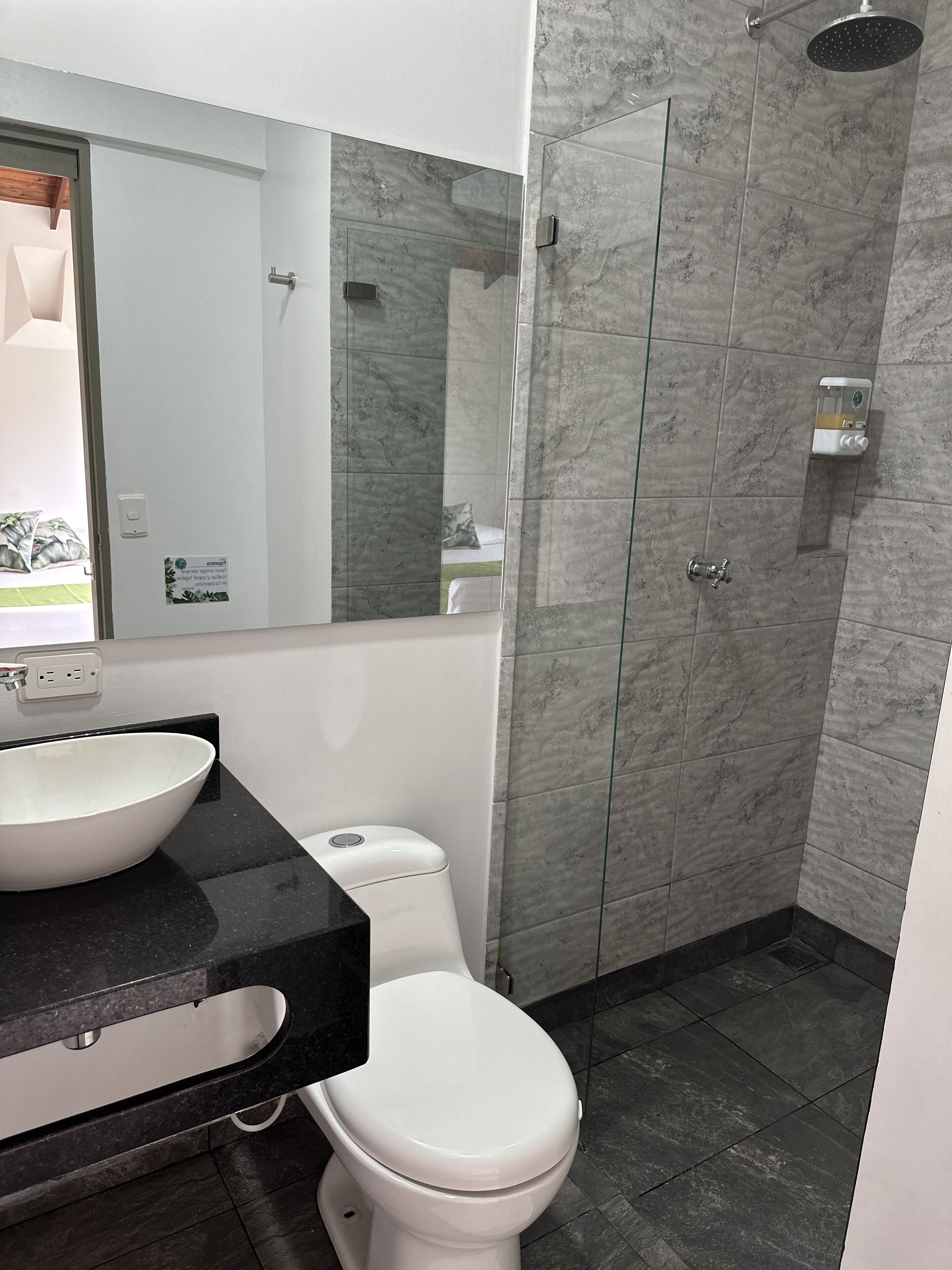 Un baño moderno con un inodoro blanco, un espejo grande, una encimera negra y un box de ducha de vidrio.