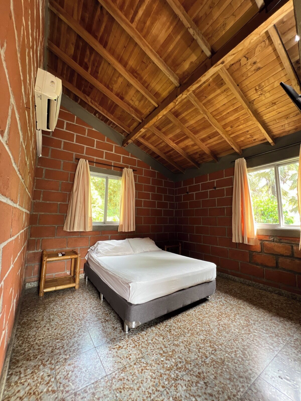 A room with a bed, a small table, a window with curtains, and a brick wall.