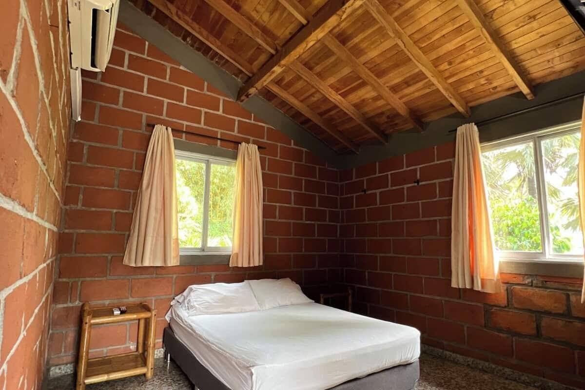 Een kamer met een bed, een klein tafeltje, een raam met gordijnen en een bakstenen muur.