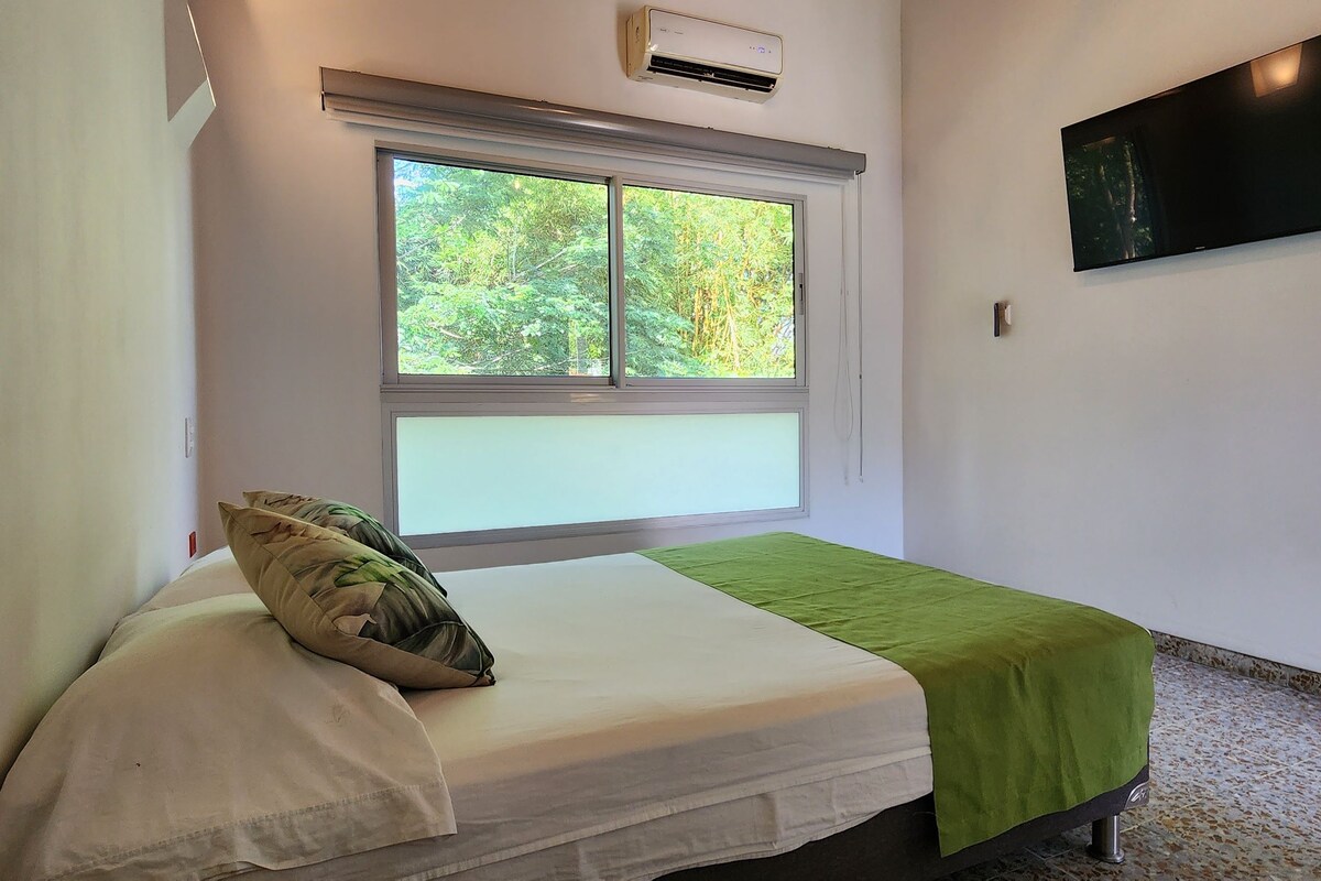 Een eenpersoonsbed met een groene sprei, een raam met uitzicht op bomen en een wandtelevisie.