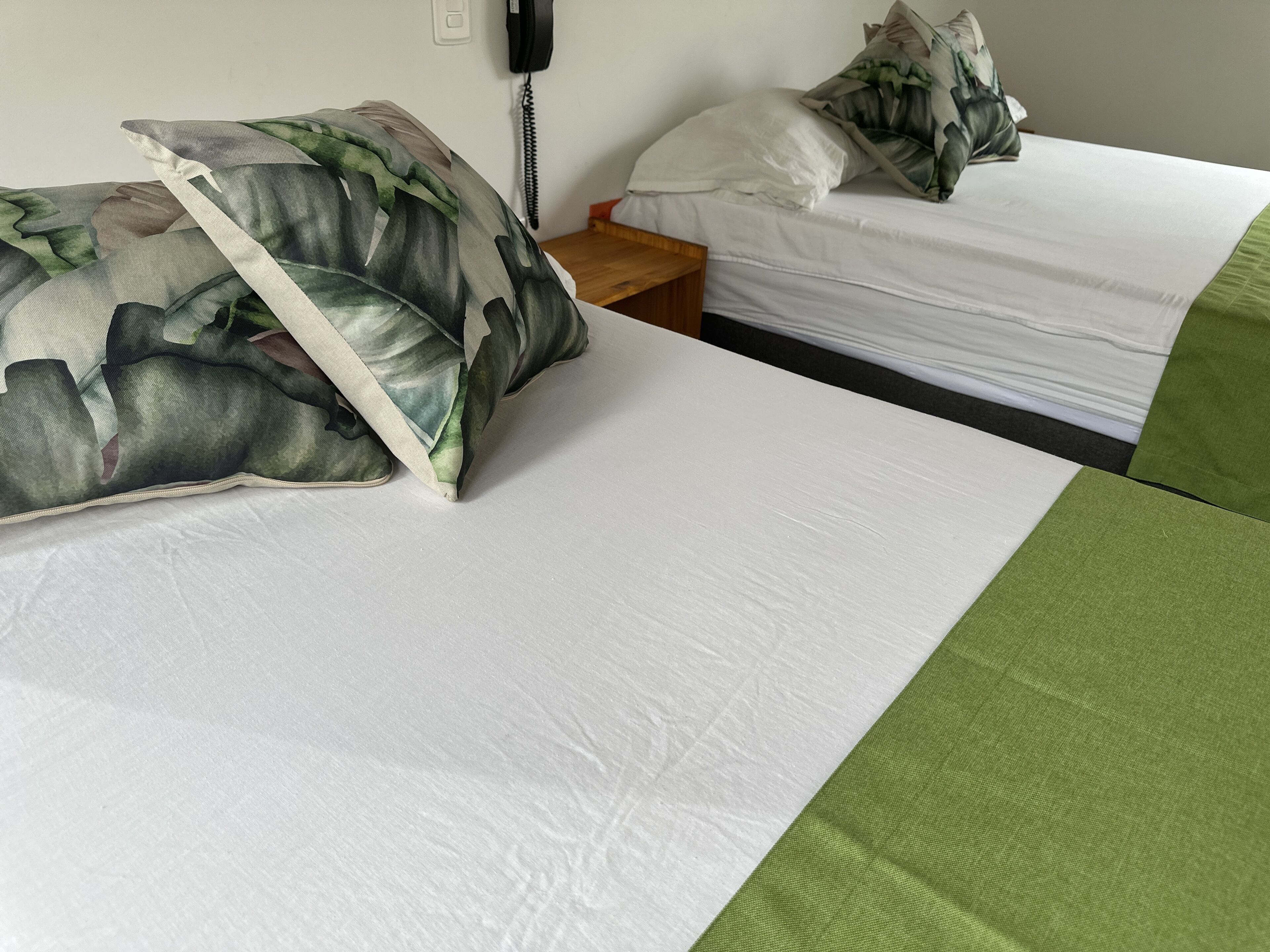 Une chambre d’hôtel avec deux lits, chacun doté d’un matelas blanc et d’un oreiller à motifs verts et blancs. Une table de chevet en bois se trouve entre les deux lits.