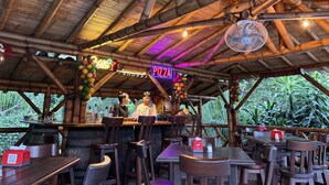 Un restaurante al aire libre con estructuras de bambú, letreros de neón que indican 'Bar' y 'Pizza', y un ventilador colgado del techo.