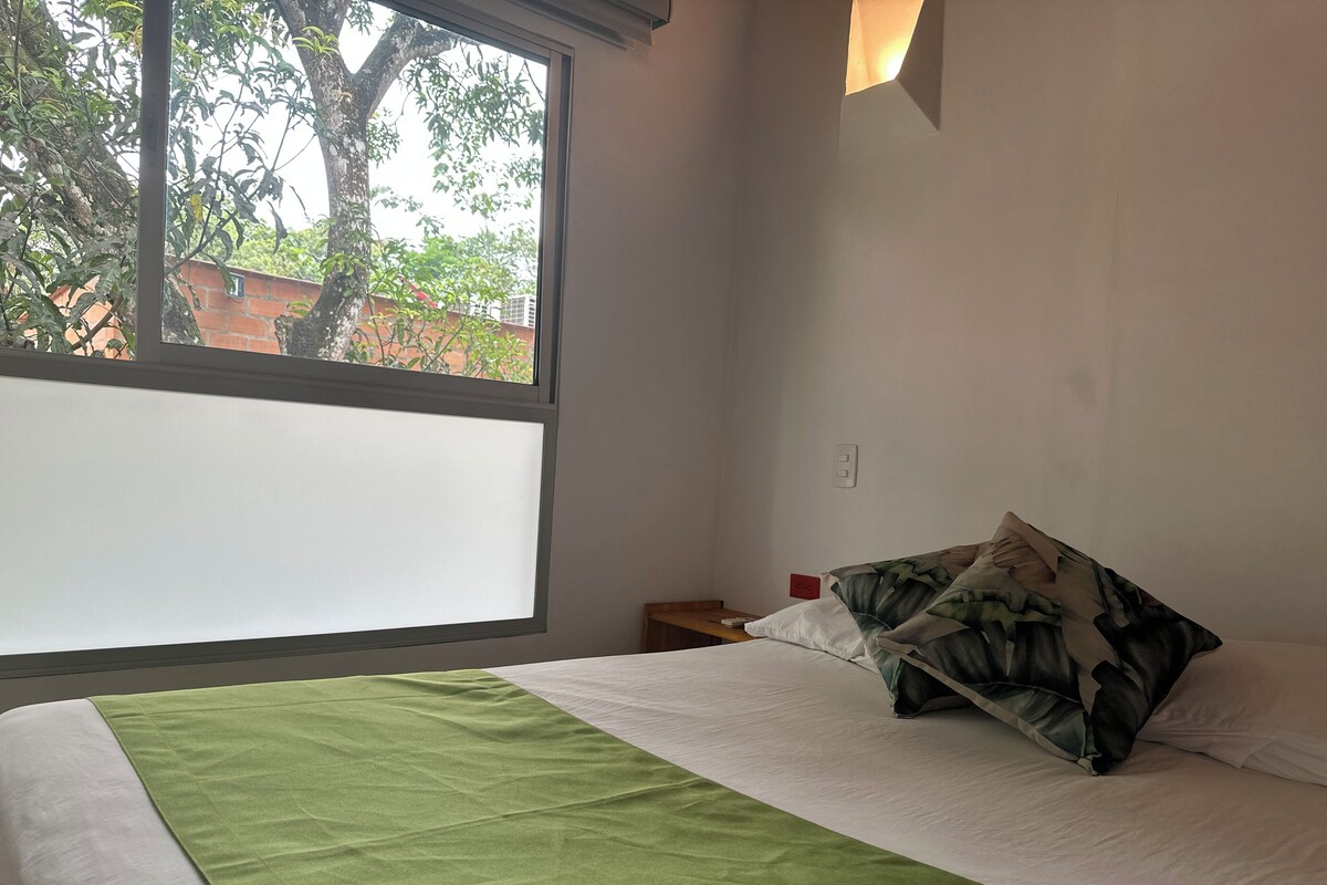 Ein Hotelzimmer mit Bett, Nachttisch, Fenster und Blick auf Bäume und Gebäude.
