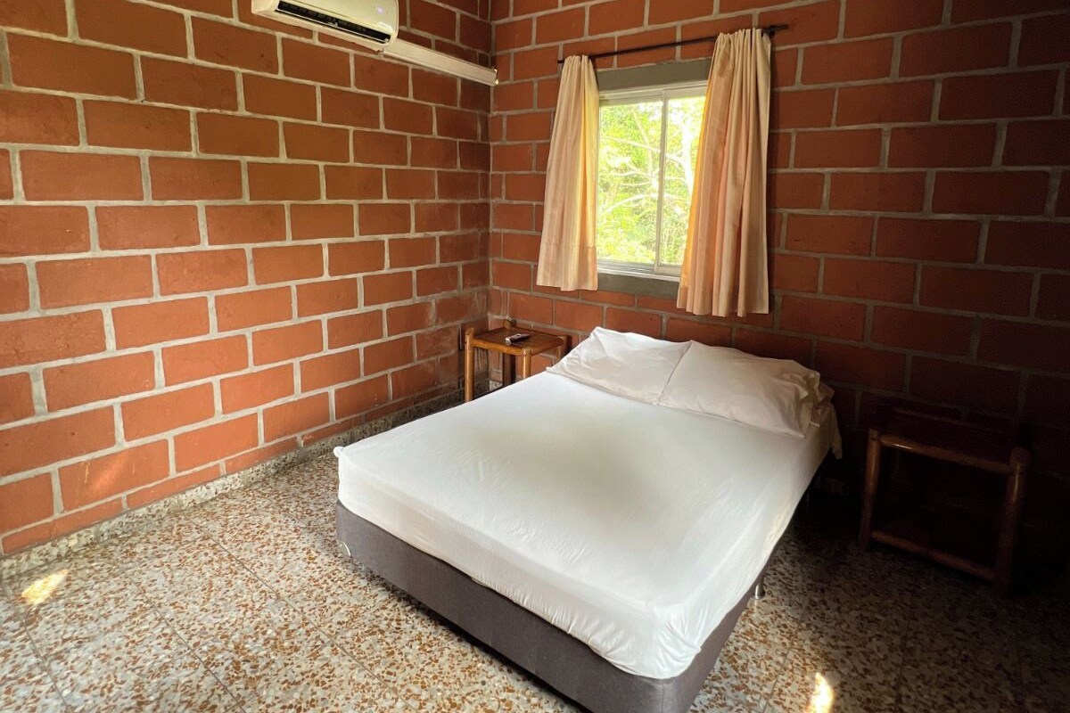A room with a brick wall, a bed, a window with curtains, and an air conditioning unit.