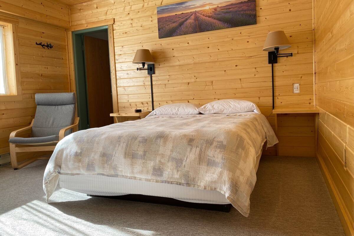 A wooden room with a bed, two nightstands, a chair, and a painting on the wall.