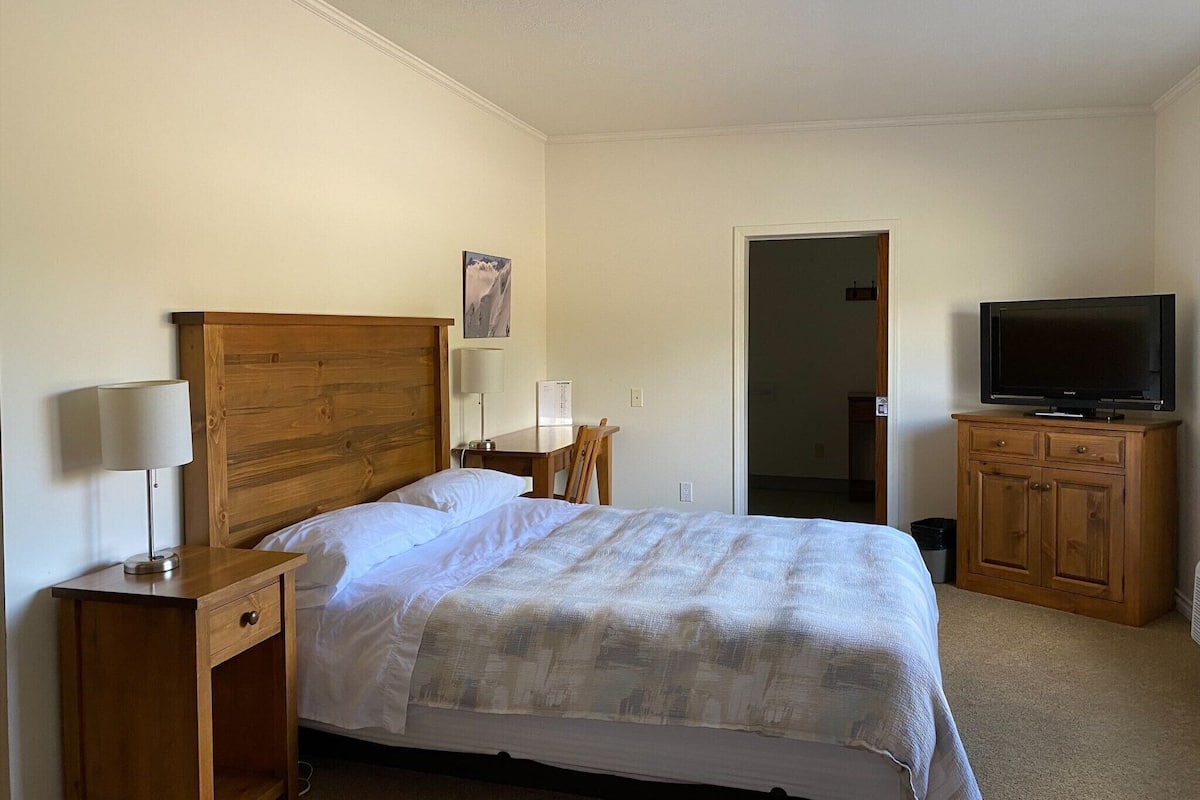A hotel room with a wooden bed, bedside lamps, a television on a wooden cabinet, and a door leading to another room.