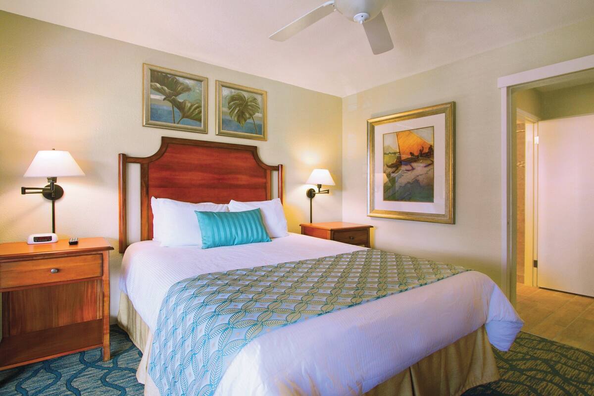 Ein Hotelzimmer mit einem großen Bett, zwei Nachttischen, einem Deckenventilator und Kunstwerken an den Wänden.