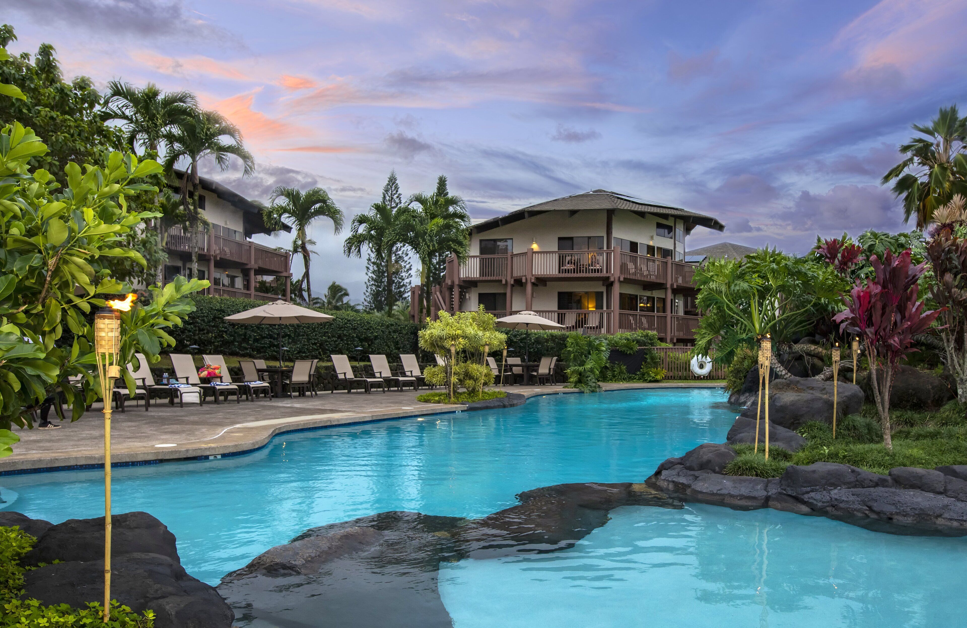 A resort pool area with lounge chairs, a large building, and tropical landscaping.