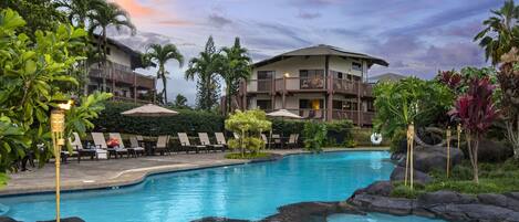 A resort pool area with lounge chairs, a large building, and tropical landscaping.