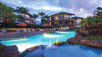 A pool area with lounge chairs and umbrellas, surrounded by rock formations and palm trees, with a building in the background.