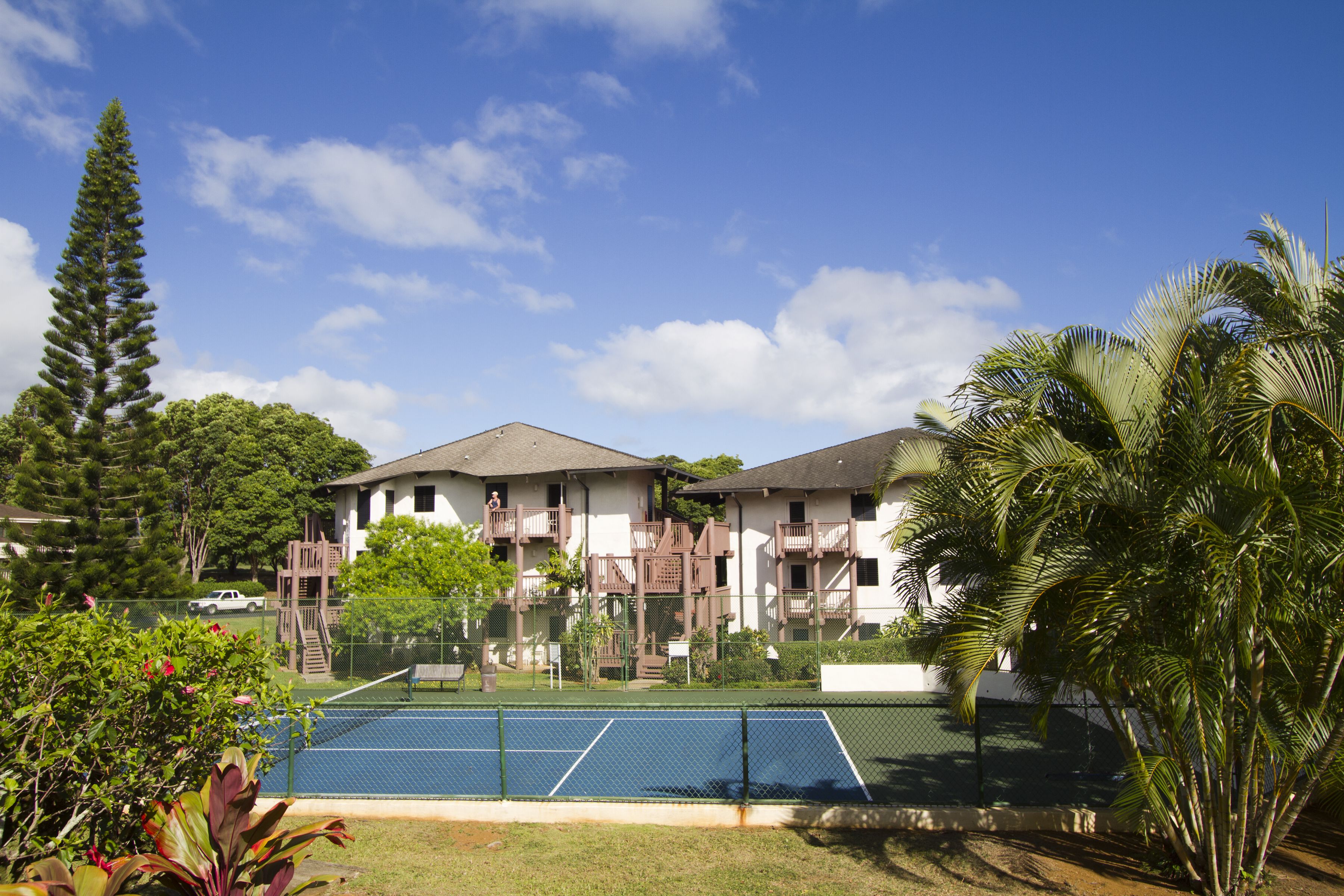 A tennis court with a net, surrounded by a fence, and adjacent to a building with balconies and a staircase.