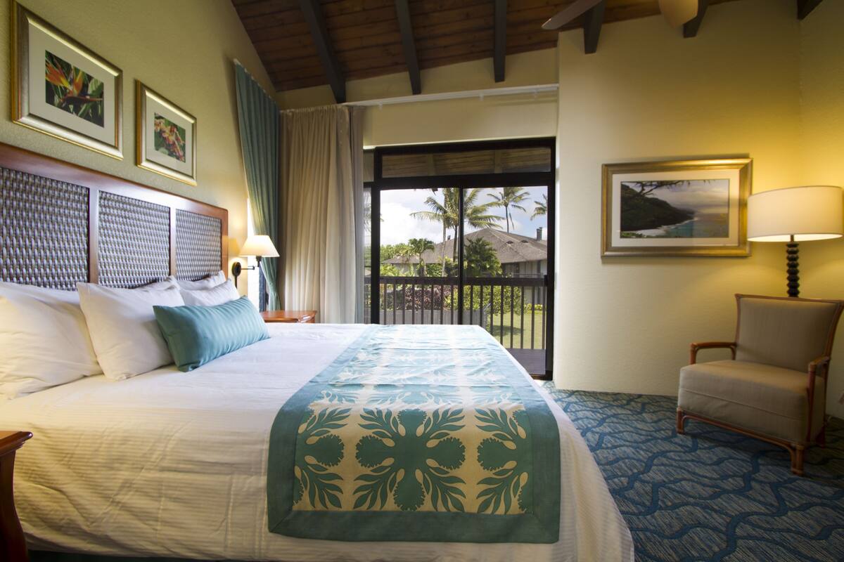Ein Hotelzimmer mit einem großen Bett, einem Sessel, einem Schreibtisch und Blick auf Palmen und einen Strand.