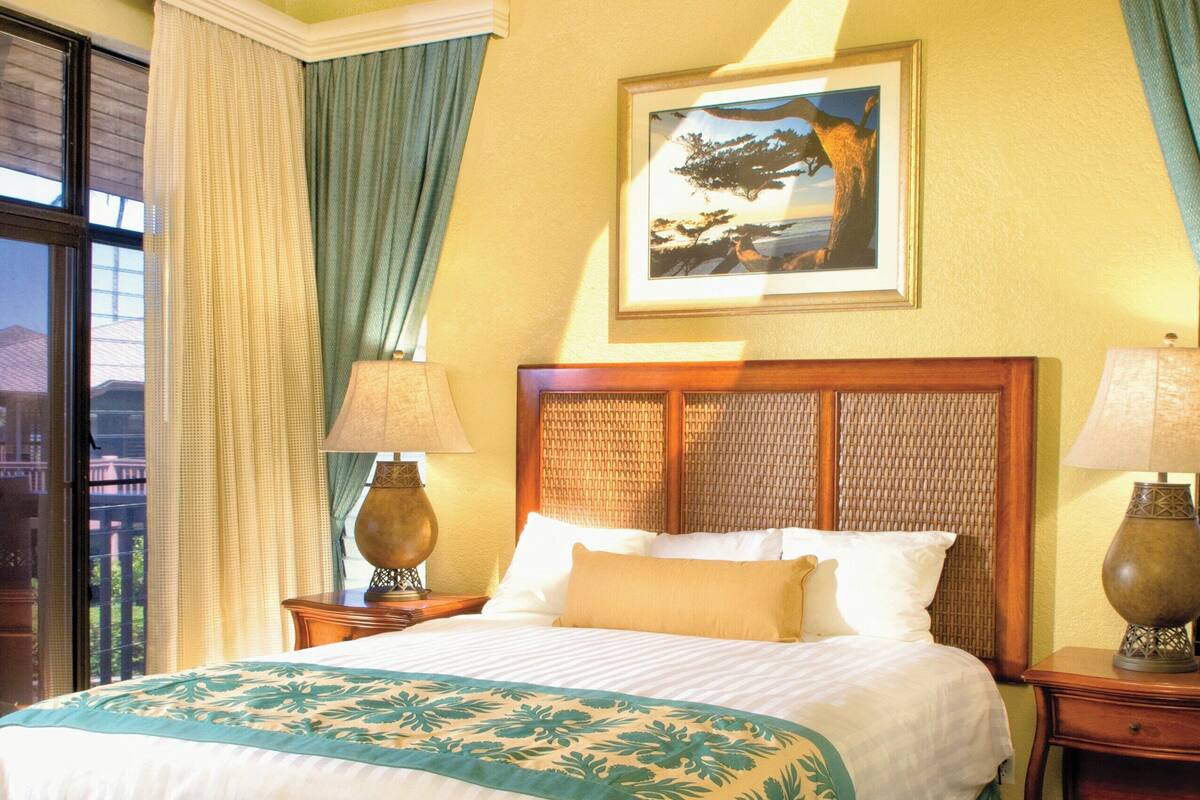 Ein Hotelzimmer mit einem großen Bett, einem Deckenventilator, einem Fenster mit Vorhängen, einem gerahmten Bild an der Wand und einem gemusterten Teppich.