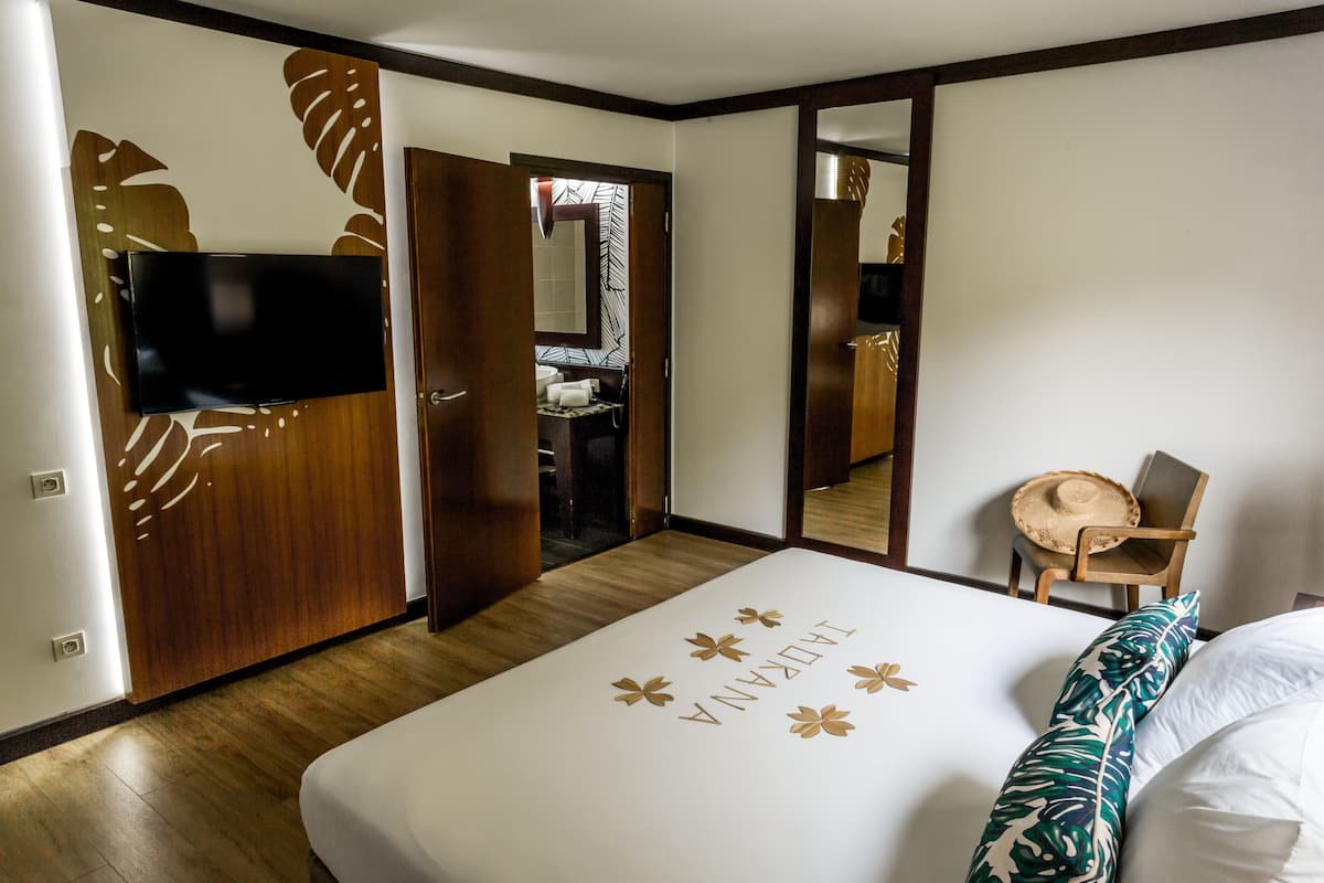 Ein Hotelzimmer mit einem Bett, einem an einer Holzvertäfelung befestigten Fernseher, einem Spiegel, einem Sessel und einem durch eine geöffnete Tür einsehbaren Badezimmer.
