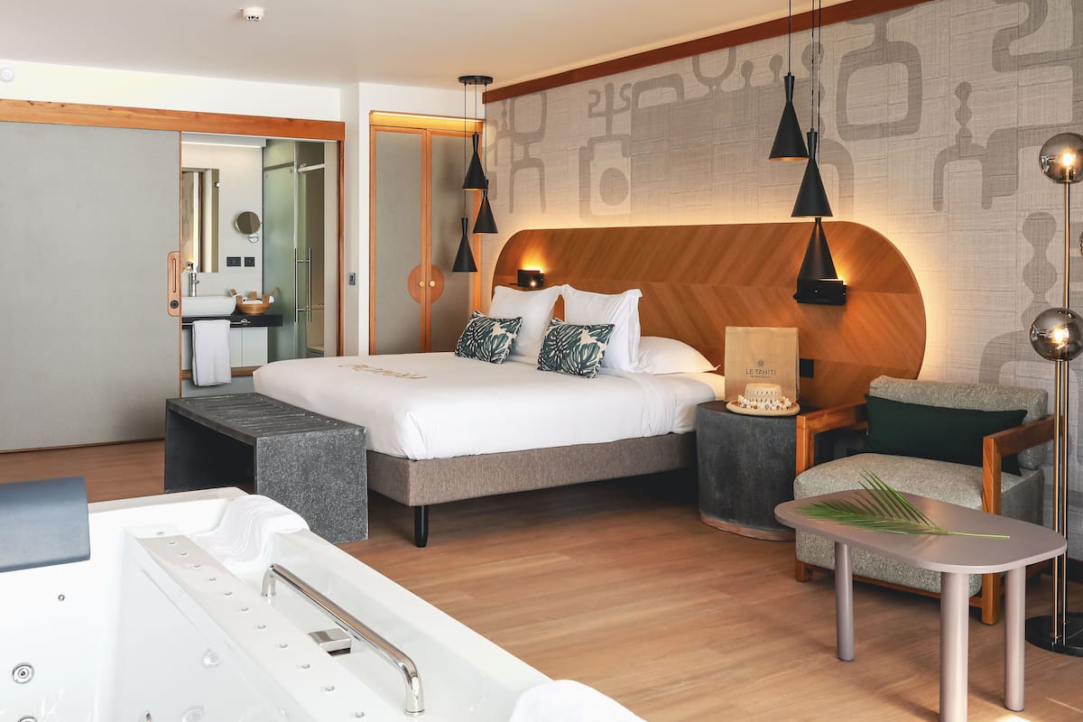 Ein modernes Hotelzimmer mit einem großen Bett, einer Sitzecke mit kleinem Tisch, einer Badewanne und einem großen Spiegel.