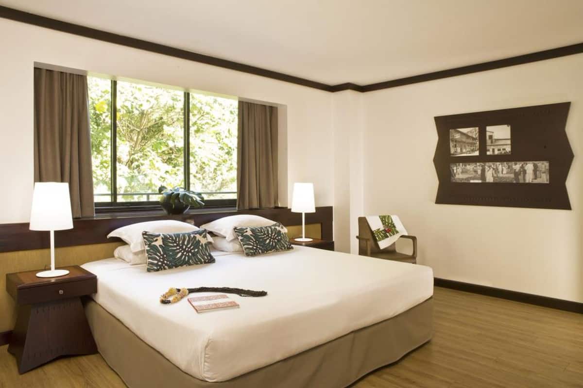 Ein Hotelzimmer mit einem großen Bett, zwei Nachttischen, einem Fenster mit Vorhängen und einem an der Wand befestigten Bilderrahmen.
