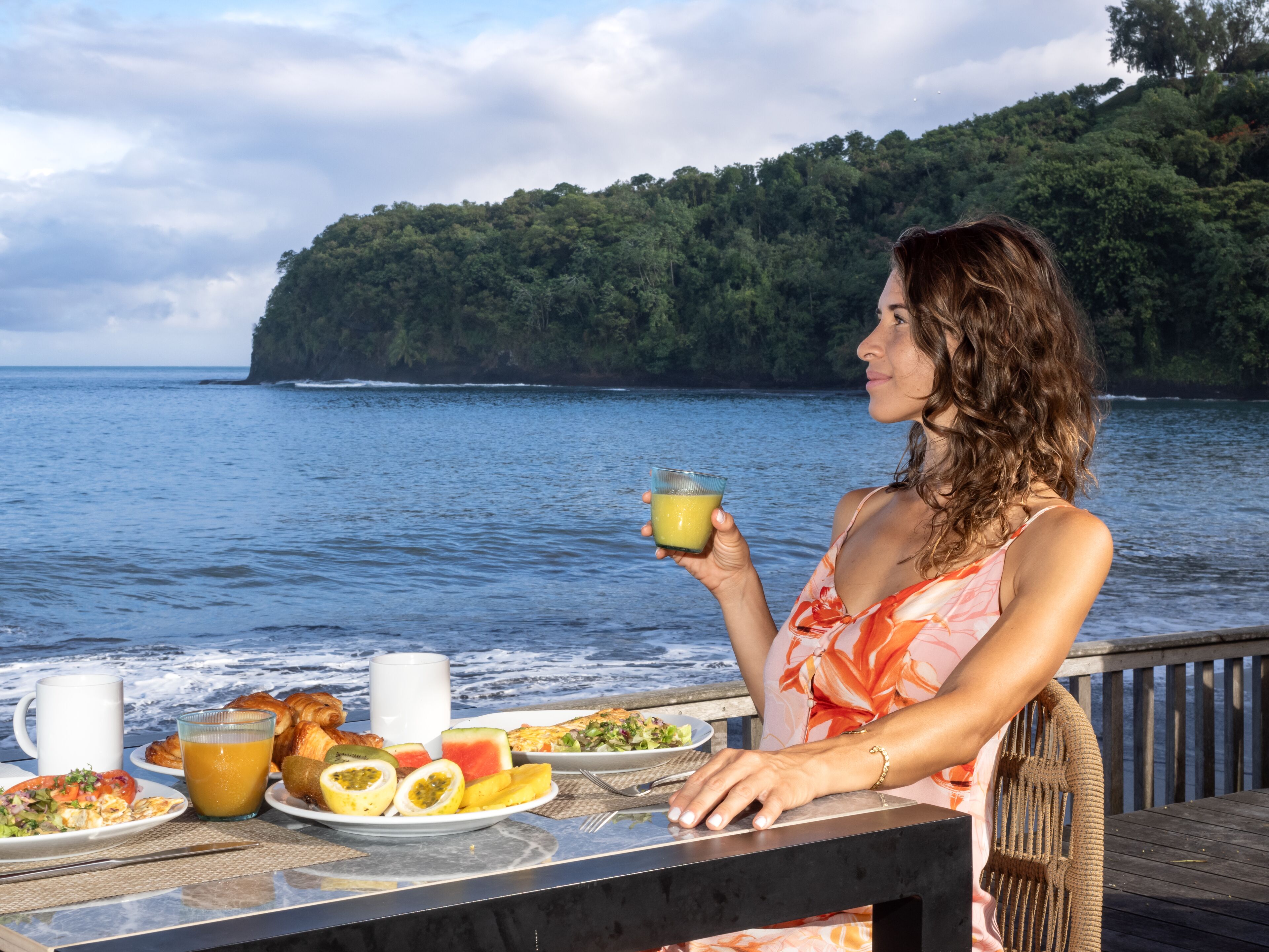 A person sitting at a table with a meal and a drink, overlooking a body of water and a forested hill.