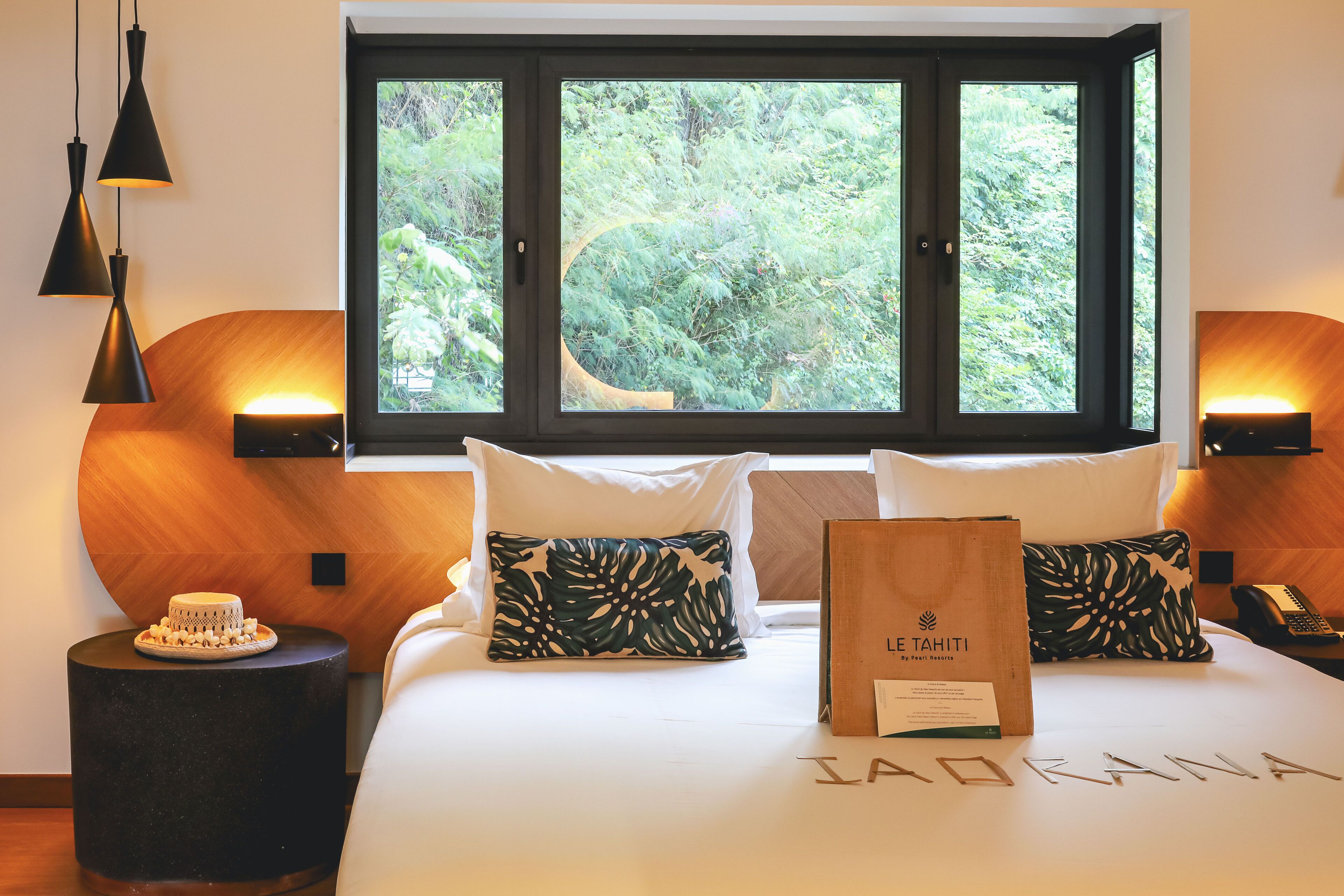Una habitación de hotel con una cama, una ventana que da a zonas verdes, un cabecero de madera y una mesita de noche con un pastel.