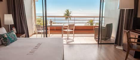 A hotel room with a bed, a desk, a chair, a balcony with a view of the beach and ocean.