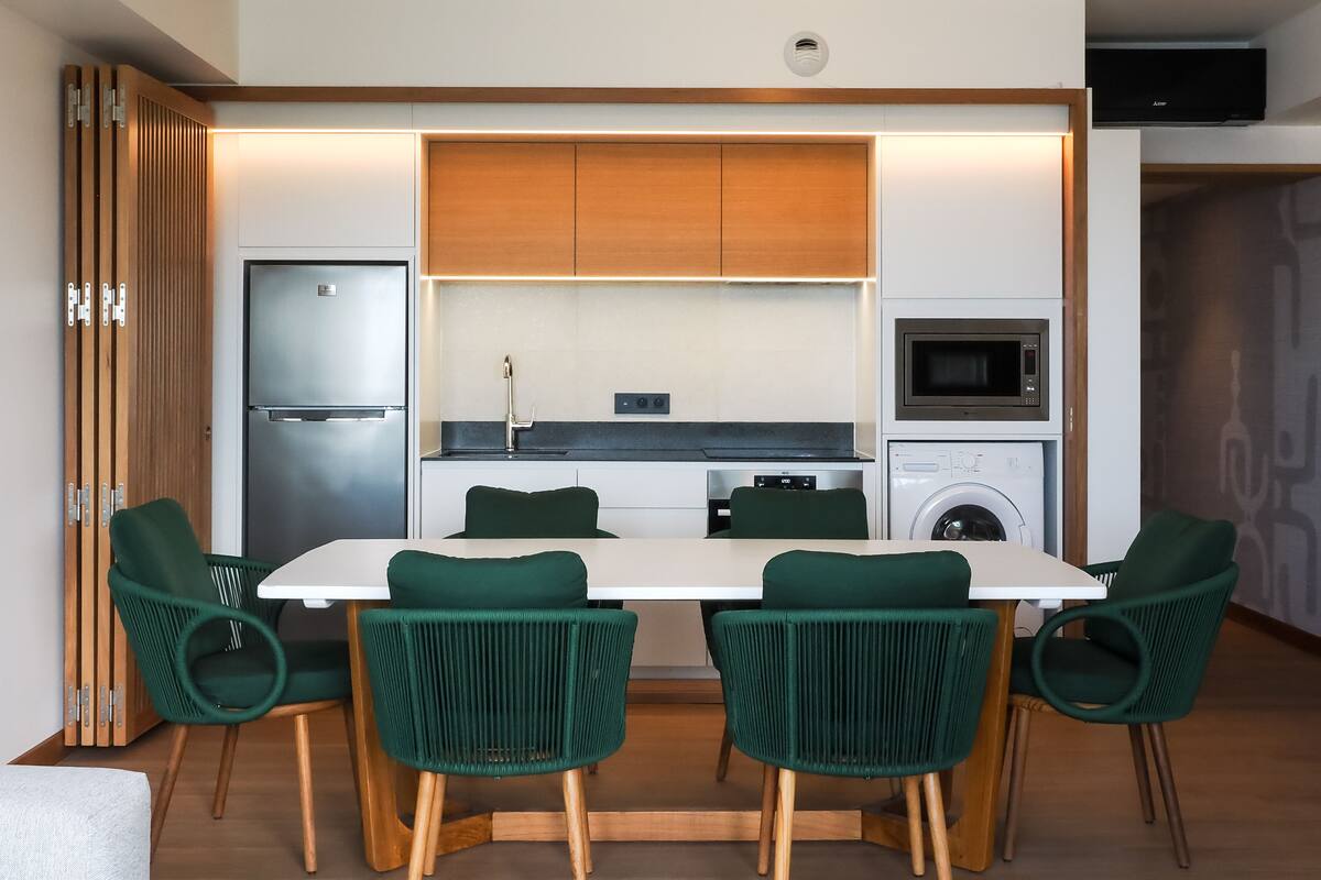 Eine moderne Küche mit Einbaukühlschrank, Mikrowelle und Waschmaschine. Ein Esstisch bietet Platz für vier grüne Stühle.