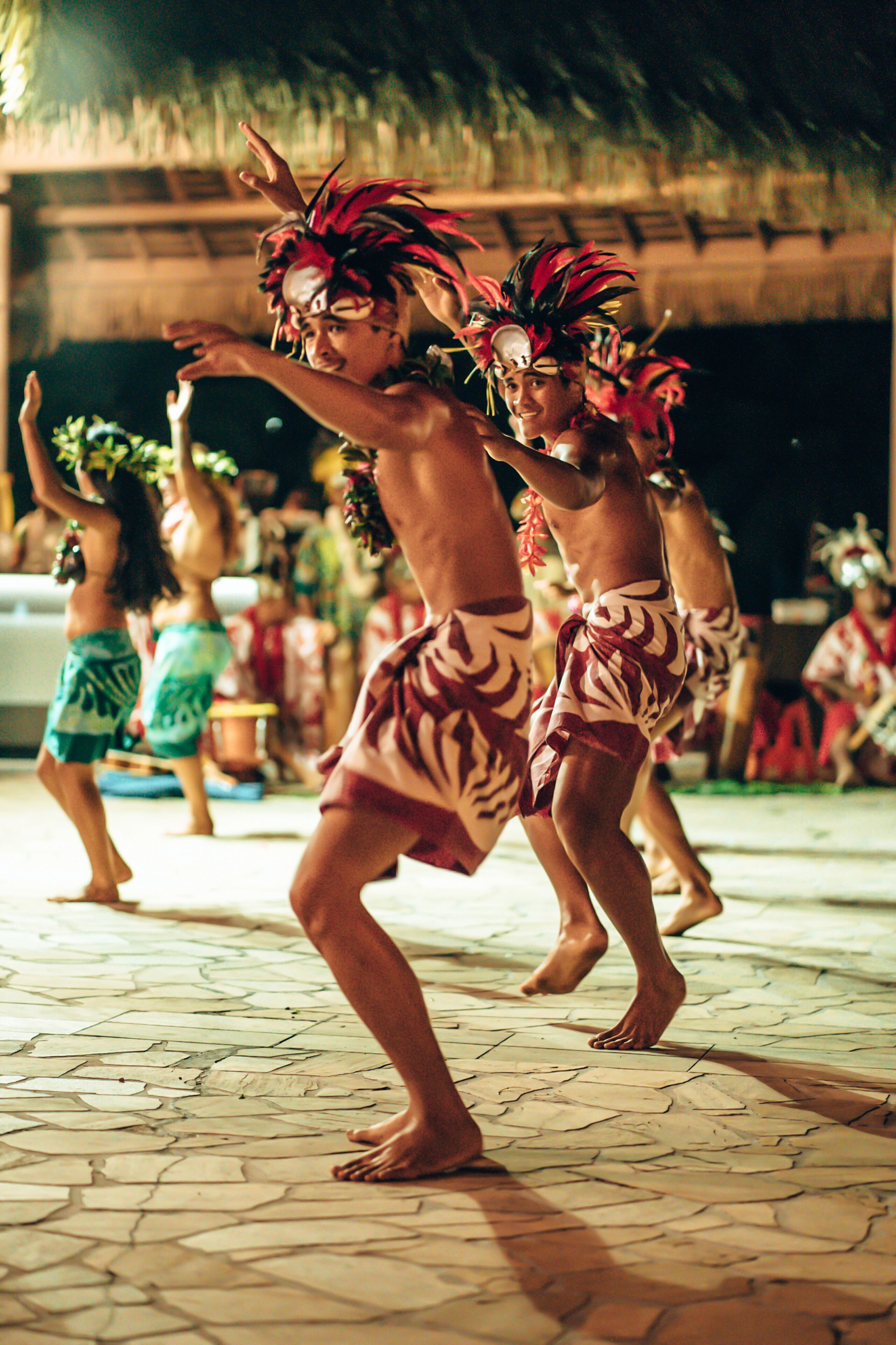 A group of people in traditional attire performing a dance on a tiled floor with a thatched roof in the background.