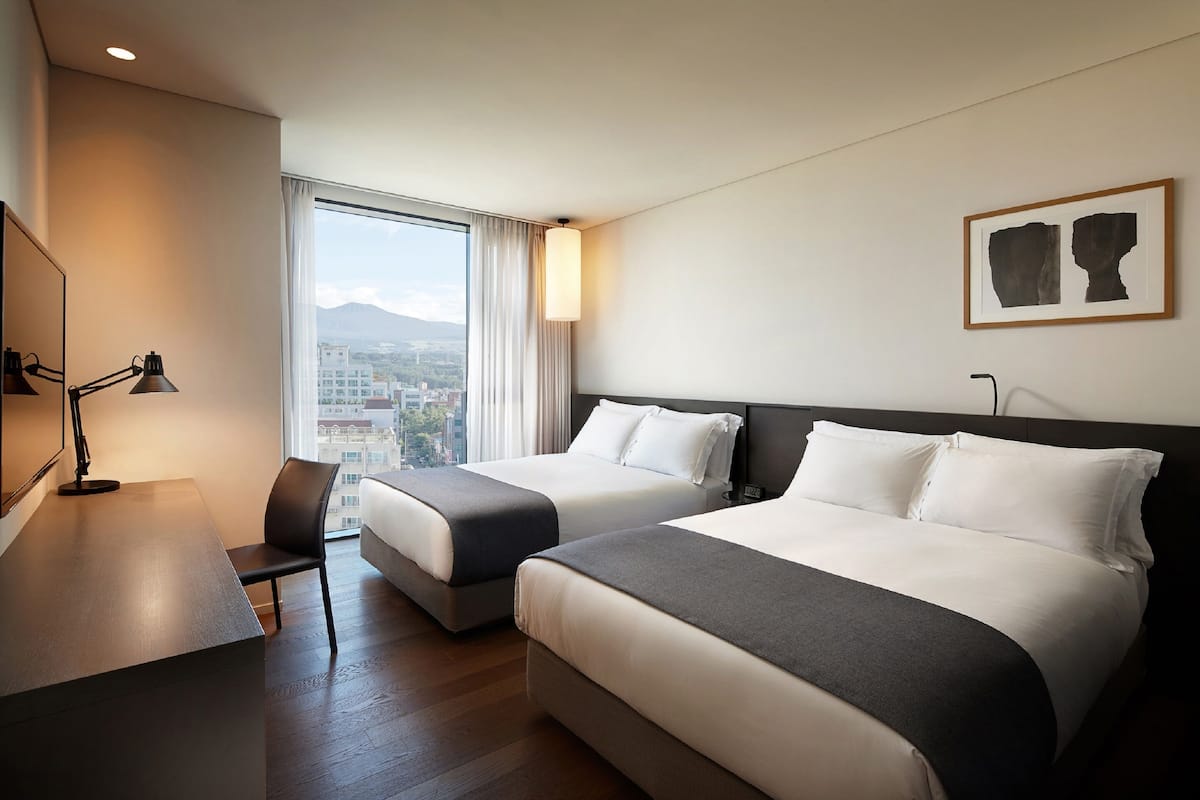 Habitación de hotel con dos camas, un escritorio, una silla, una lámpara y un ventanal con vistas a edificios y montañas.
